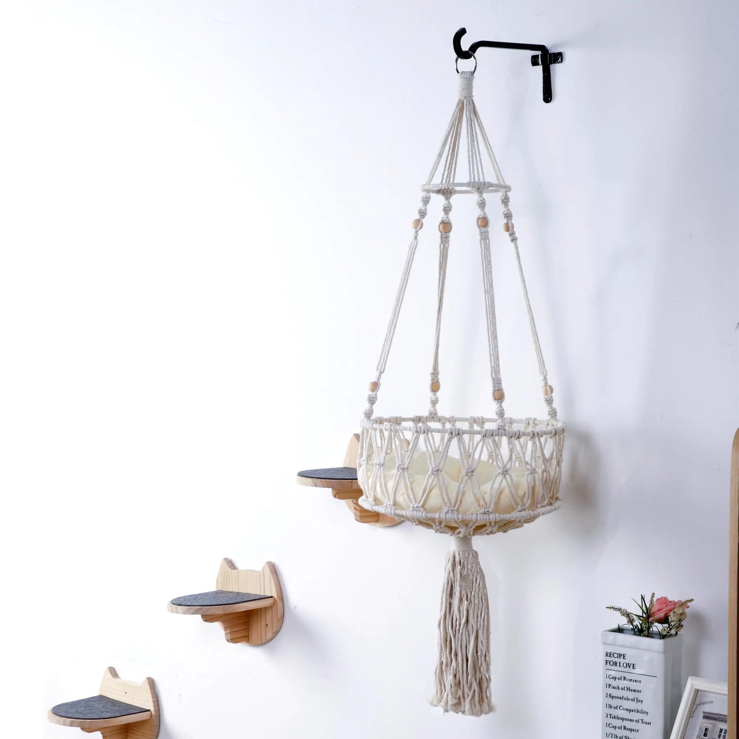 Macrame cat hammock with cat shelves and tassels hanging on a white wall.