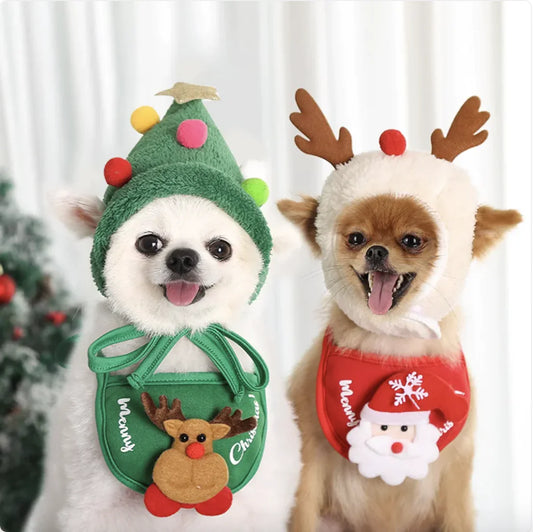 Two small dogs wearing Christmas-themed outfits with a green tree design pet hat and a reindeer pet bib, and a red outfit with Santa Claus design pet bib and a white reindeer pet hat, against a white curtain background.