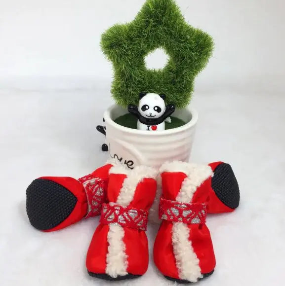 Red dog boots with white fur trim on a white background, next to a small potted plant with a panda figurine.
